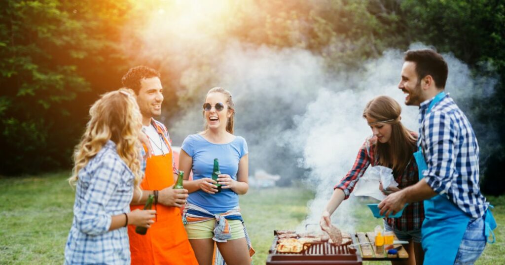 Grupa młodych ludzi świętuje w ogrodzie przy grillu, w radosnej atmosferze z piwem w ręku, w otoczeniu zieleni i dymu z rusztu – Odliczanie do Majówki.